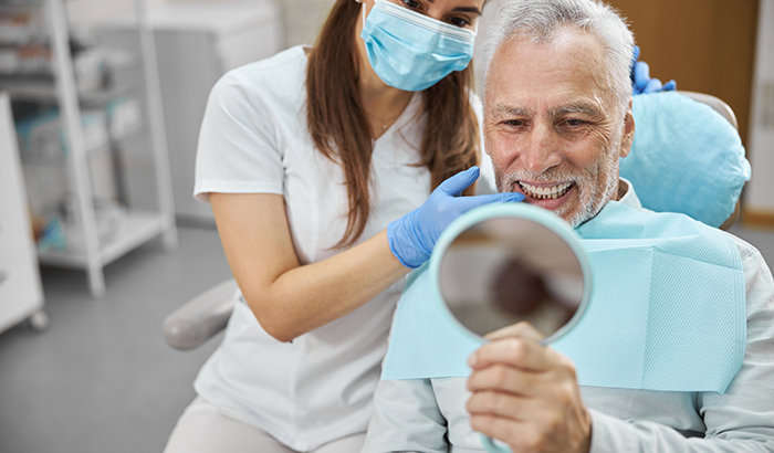 Older man in a dental chair holding up a hand mirror and looking at his cosmetic dentistry from Encinitas Periodontics & Dental Implants in Encinitas, CA